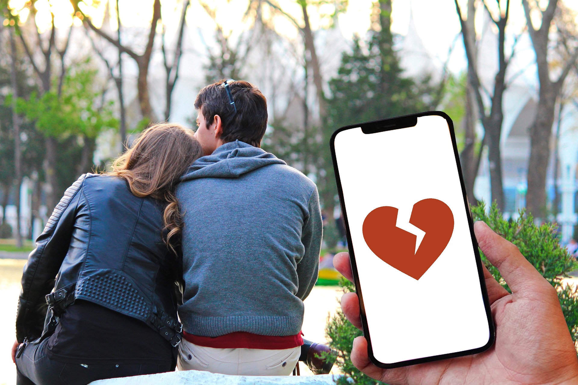 Um casal ao fundo da imagem e em primeiro plano um celular com a imagem de um coração partido