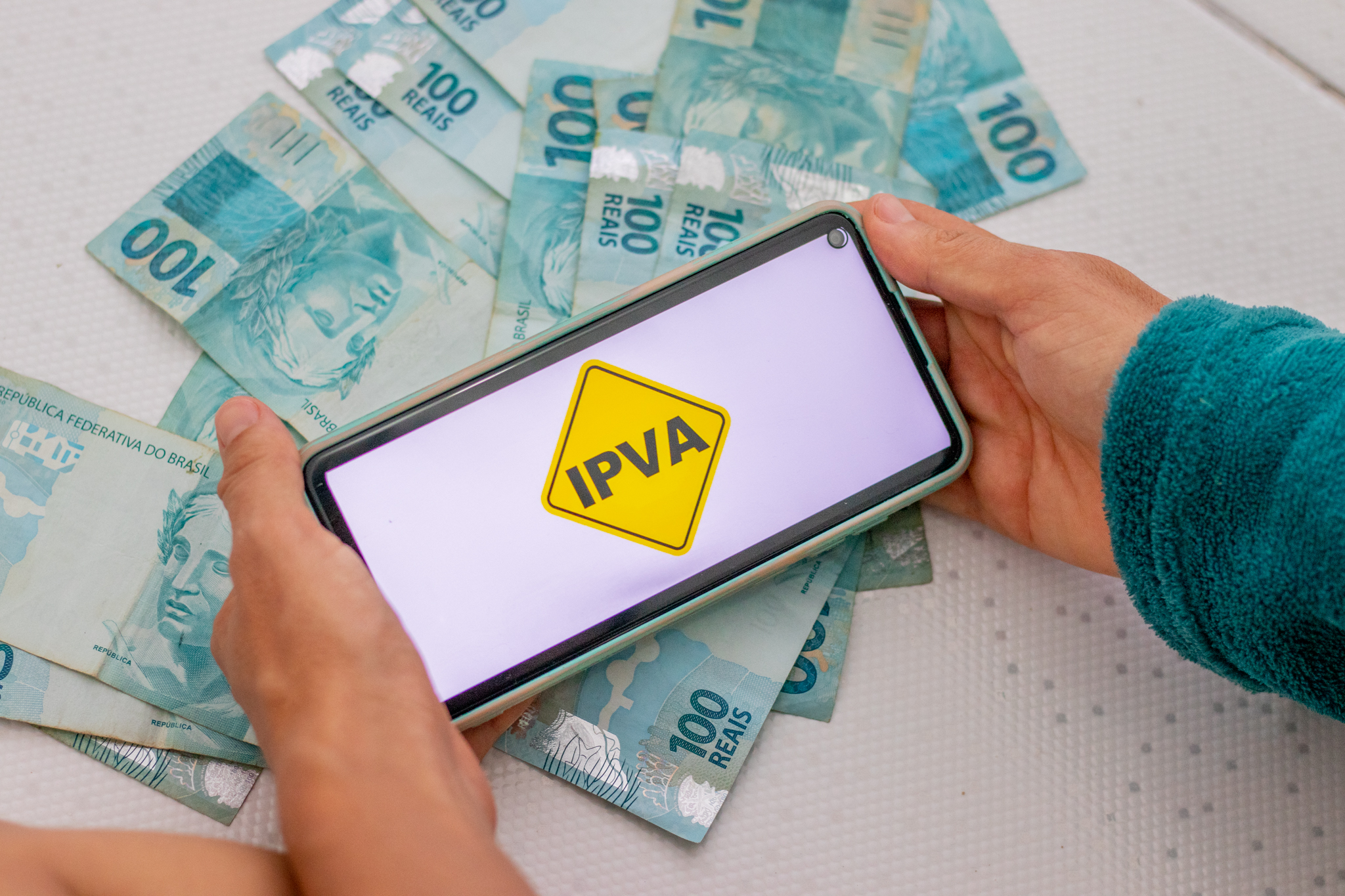 Cédulas de dinheiro brasileiro e uma pessoa segurando um celular com o papel de parede escrito 'ipva'