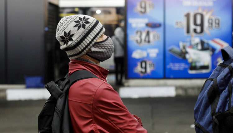 A imagem mostra uma pessoa de perfil, usando um gorro com padrões em preto e branco e uma máscara facial. A pessoa está vestindo uma jaqueta vermelha e parece estar em um ambiente urbano, com um fundo que apresenta um cartaz publicitário com preços e produtos. O ambiente é iluminado e parece ser uma área de comércio.
