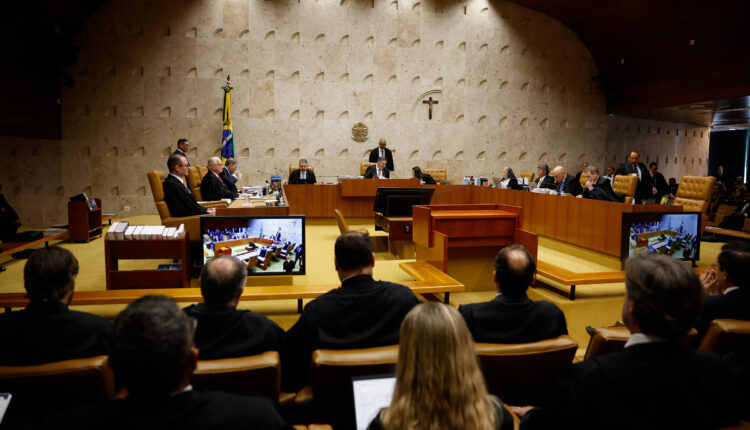 A imagem mostra uma sessão do Supremo Tribunal Federal do Brasil. No centro, há uma mesa com vários juízes e um presidente da sessão, todos em trajes formais. Ao fundo, há uma bandeira do Brasil e um crucifixo na parede. Na frente, há uma plateia com pessoas sentadas, algumas delas com trajes escuros. Em duas telas de projeção, é possível ver informações relacionadas à sessão.