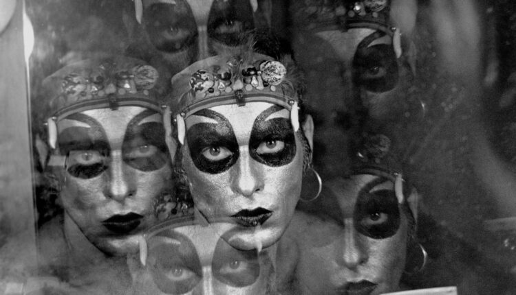 A imagem apresenta um retrato em preto e branco de uma pessoa com maquiagem dramática, incluindo olhos destacados em preto e lábios escuros. A figura parece refletida em uma superfície, criando um efeito de múltiplas camadas. O fundo é desfocado, e há um leve brilho que sugere um ambiente de backstage ou de performance.
