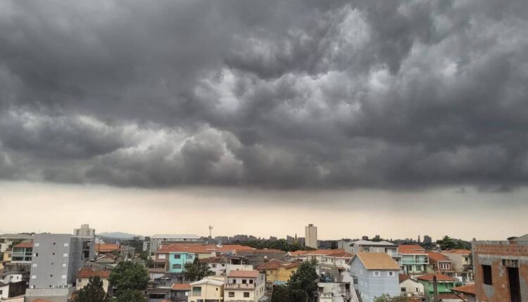 A imagem mostra uma vista panorâmica de uma cidade sob um céu nublado e escuro. As nuvens são densas e ameaçadoras, cobrindo a maior parte do céu. Abaixo, pode-se ver uma variedade de edifícios e casas com telhados de diferentes cores, predominantemente em tons de terracota e cinza. O horizonte é visível ao fundo, com algumas estruturas mais altas se destacando.