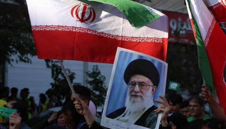 A imagem mostra um grupo de pessoas em um protesto, segurando bandeiras do Irã. Algumas pessoas também estão segurando um retrato de um líder iraniano. O ambiente parece ser urbano, com edifícios ao fundo e uma atmosfera de manifestação.