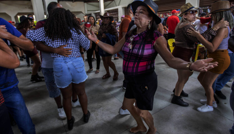 A imagem mostra um grupo de pessoas dançando em um ambiente festivo, possivelmente durante uma festa junina. No centro, uma mulher de cabelo escuro e encaracolado, vestindo uma blusa listrada e uma saia preta, dança com os braços abertos. Ao fundo, outras pessoas também dançam, algumas em pares, e estão vestidas com roupas típicas de festa junina, como camisas xadrez e chapéus de palha. O ambiente é iluminado e parece ser um espaço amplo.