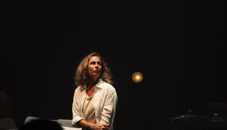 Uma mulher com cabelo cacheado e solto, vestindo uma camisa branca, está em um palco com iluminação suave. Ela parece estar em um momento de reflexão ou emoção, olhando para o lado. O fundo é escuro, com uma luz amarelada visível ao fundo, e há alguns objetos de cena, como cadeiras, em um ambiente teatral.