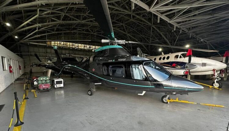 A imagem mostra um helicóptero preto com detalhes em verde, estacionado em um hangar. O helicóptero está em primeiro plano, com suas lâminas visíveis. Ao fundo, há um avião pequeno e outros equipamentos de aviação. O piso do hangar é de concreto e há algumas luzes acesas no teto.