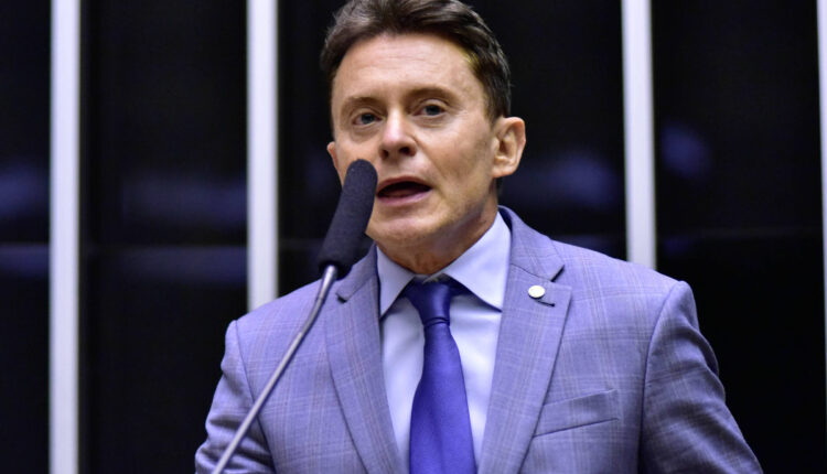 Um homem em um terno cinza claro e gravata azul está falando em um microfone em um ambiente de parlamento. Ele parece estar em uma posição de destaque, com um fundo que inclui painéis verticais. O homem tem cabelo castanho e está expressando-se de forma enérgica.