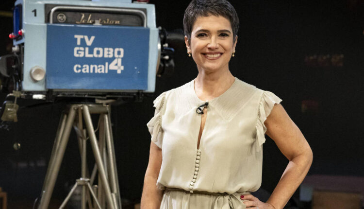 Uma mulher sorridente está em pé em um estúdio de televisão, vestindo um vestido claro. Ao fundo, há uma câmera de TV antiga com a inscrição