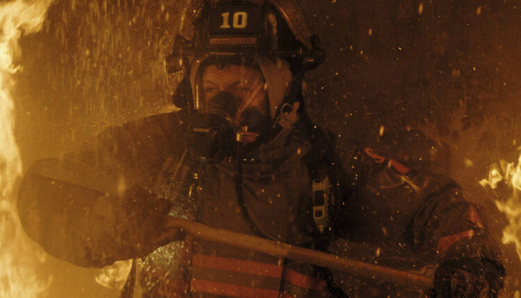 Um bombeiro está lutando contra um incêndio em um ambiente cheio de chamas e fumaça. Ele usa um capacete com o número 10 e uma máscara de proteção, segurando uma ferramenta de combate a incêndios. O fundo é dominado por fogo e fumaça, criando uma atmosfera intensa e perigosa.