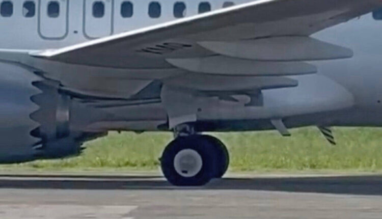 A imagem mostra a parte inferior de um avião, focando na roda do trem de pouso. O avião é de cor cinza e está posicionado em uma pista de pouso, com grama verde ao fundo. As asas do avião são visíveis, assim como parte do motor.