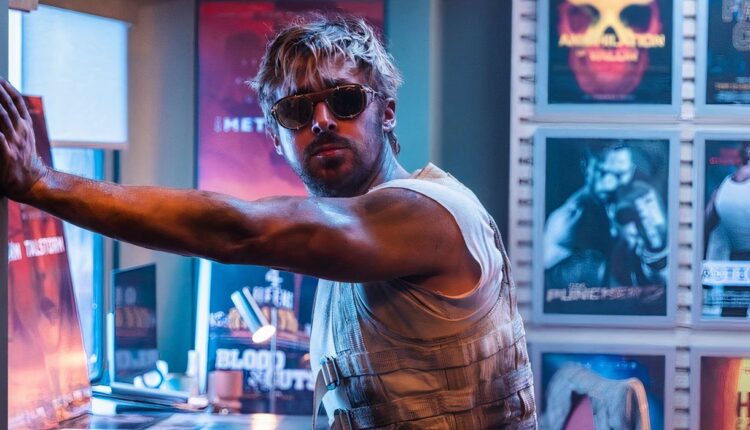 Ryan Gosling wearing sunglasses and a dirty vest stands in a room full of movie posters in The Fall Guy.