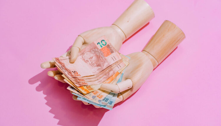 A imagem mostra um par de mãos de boneca segurando várias notas de dinheiro em cima de um fundo rosa. As notas são de diferentes valores e cores, destacando-se na composição.