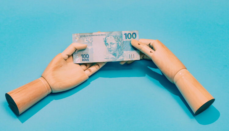 A imagem mostra duas mãos de madeira, uma de cada lado, segurando uma nota de 100 reais. O fundo é azul claro, destacando a nota que possui a figura de uma mulher e o valor impresso em destaque. As mãos estão posicionadas de forma a parecer que estão trocando a nota.