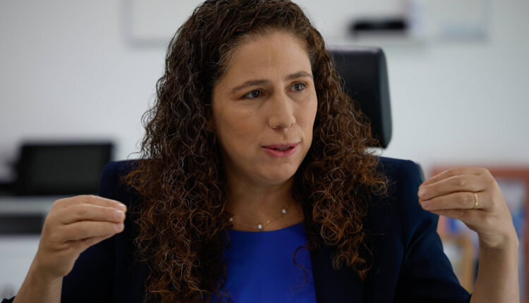 Uma mulher com cabelo cacheado e castanho claro está sentada em uma cadeira, gesticulando com as mãos enquanto fala. Ela usa uma blusa azul e um blazer escuro. O fundo é um ambiente de escritório, com uma mesa e um computador visíveis. A mulher parece estar em um momento de discussão ou entrevista.
