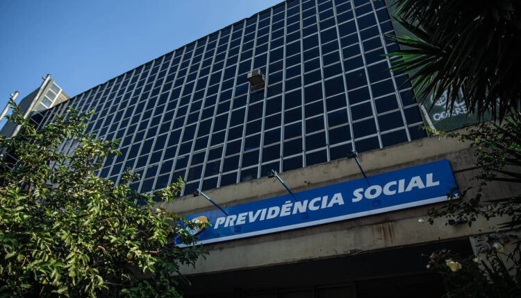 A imagem mostra a entrada de um edifício com uma fachada de vidro. Acima da entrada, há uma placa azul com o texto