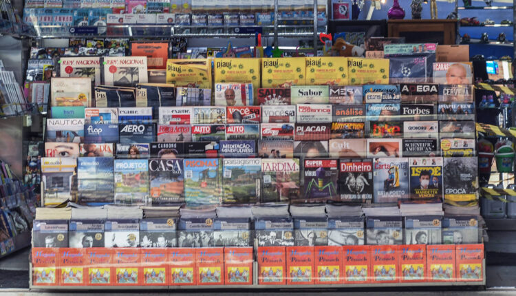 A imagem mostra uma banca de jornal com uma variedade de revistas e jornais expostos. A banca está organizada em prateleiras, com capas visíveis de diferentes publicações. À direita, uma mulher está de pé, usando uma máscara facial. O ambiente é iluminado e a banca é coberta por uma lona azul na parte superior.