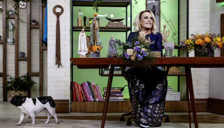 Uma apresentadora está sentada em uma mesa de madeira em um estúdio decorado. Ela usa um vestido azul escuro com detalhes em dourado e está cercada por arranjos de flores. Ao fundo, há prateleiras com livros e objetos decorativos. Um cão de pequeno porte, com pelagem branca e manchas pretas, caminha pelo chão ao lado da mesa.