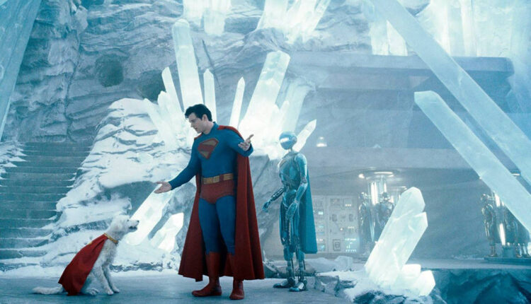 A imagem mostra um personagem vestido como super-herói, com um traje azul e uma capa vermelha, interagindo com um cachorro que também usa uma capa vermelha. Ao fundo, há uma estrutura de cristais de gelo e um robô prateado. O ambiente é gelado e iluminado, com uma atmosfera futurista.