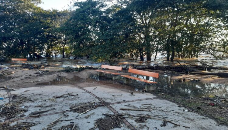 A imagem mostra uma área inundada, com água cobrindo parte do solo e detritos espalhados. Há bancos de madeira laranja visíveis, parcialmente submersos. Ao fundo, árvores estão presentes, e a luz do sol ilumina a cena, indicando que é durante o dia. A área parece ter sofrido os efeitos de uma enchente recente.