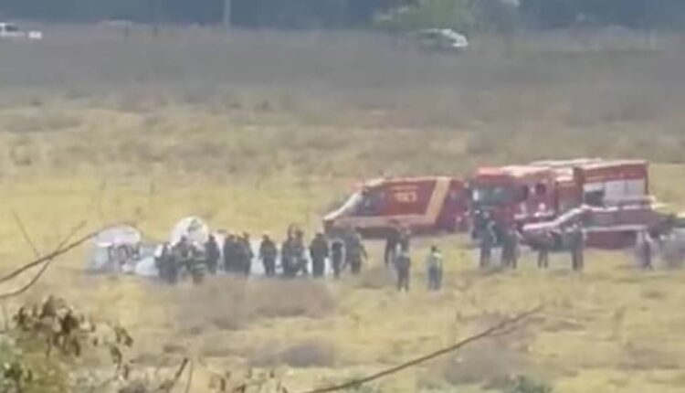 A imagem mostra um campo aberto onde ocorreu um acidente com uma aeronave. Há várias pessoas reunidas em torno do local do acidente, incluindo equipes de emergência. Ao fundo, é possível ver duas ambulâncias e um veículo de resgate. O ambiente é predominantemente seco e com vegetação baixa.