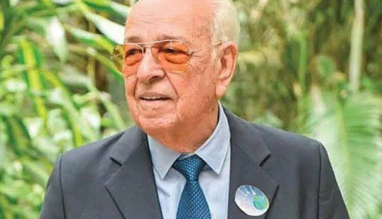 Um homem idoso está posando em um ambiente natural, cercado por plantas verdes. Ele usa um terno escuro, uma camisa clara e óculos de sol laranja. Na lapela do seu paletó, há um botão com um desenho que parece representar o planeta Terra.