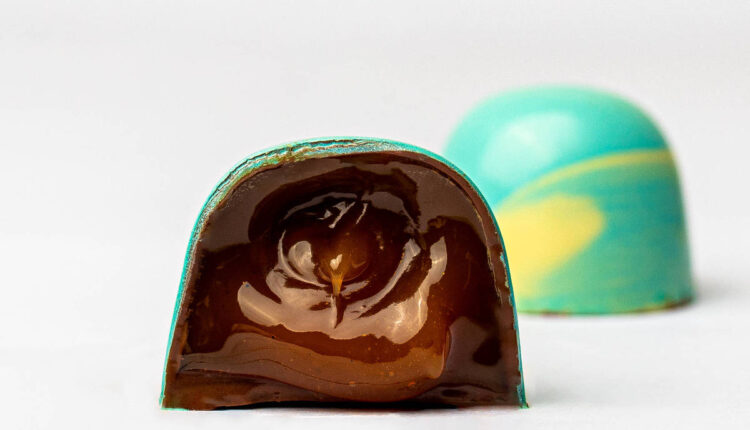 A imagem mostra um bombom de chocolate com uma casca colorida em azul e amarelo. O bombom está cortado ao meio, revelando um recheio de chocolate escuro e brilhante. Ao fundo, há outro bombom inteiro, também com a casca colorida, mas não cortado.