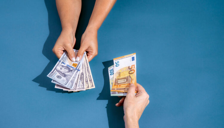 A imagem mostra duas mãos em um fundo azul. Uma mão está segurando várias notas de dólar, enquanto a outra mão está segurando uma nota de cinquenta euros. As mãos estão se aproximando, sugerindo uma troca de dinheiro.