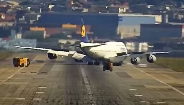 A imagem mostra um grande avião comercial, possivelmente um Boeing 747, na pista de decolagem de um aeroporto. O avião está em uma posição inclinada, com a parte traseira levantada. Ao lado do avião, há um veículo de serviço de aeroporto, que parece estar realizando alguma atividade de apoio. Ao fundo, é possível ver a cidade com prédios e uma variedade de cores nas construções.