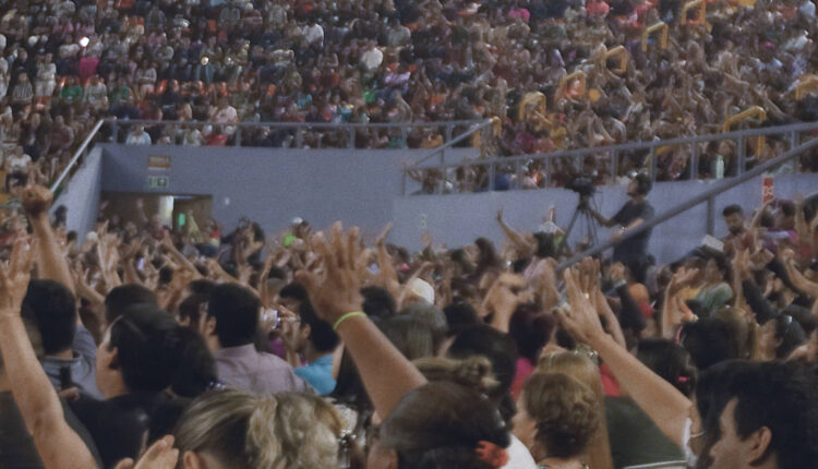 A imagem mostra uma grande multidão em um evento, com muitas pessoas levantando as mãos. O ambiente parece ser um auditório ou arena, com assentos ocupados e uma grande quantidade de espectadores visíveis. A iluminação é suave e o público parece estar engajado ou participando ativamente do que está acontecendo no palco.