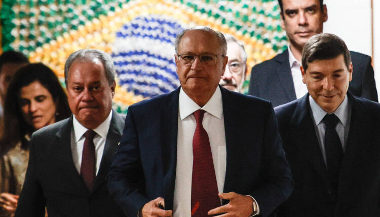 A imagem mostra um grupo de homens e mulheres em um ambiente formal. No centro, um homem de terno escuro e gravata vermelha caminha em direção à câmera, enquanto outros dois homens, um à sua esquerda e outro à direita, também estão vestidos formalmente. Ao fundo, há uma parede decorada com um padrão que remete à bandeira do Brasil, e algumas pessoas estão visíveis atrás deles. O ambiente parece ser um espaço de reunião ou evento oficial.