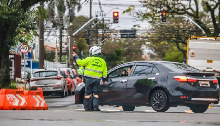 Guarda de trânsito de Curitiba orientando um carro que está passando por um trecho que tem uma rua bloqueada para obras.
